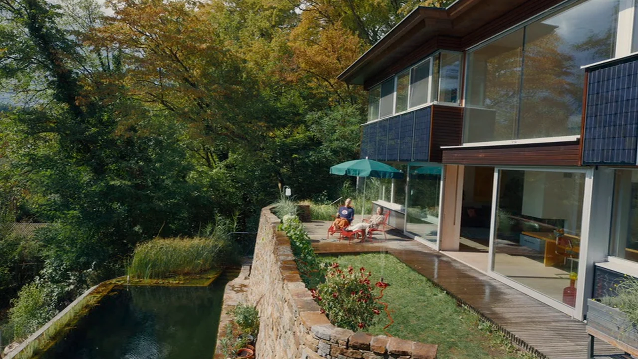 Club Zero (2023) movie still: wide — A modern multi-story glass-walled house overlooks a stone retaining wall and a pond. Two older adults sit on…