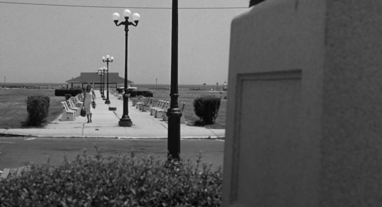 Stardust Memories (1980) movie still: wide — A seaside promenade features rows of white benches and black lamp posts under daylight. A woman in a…