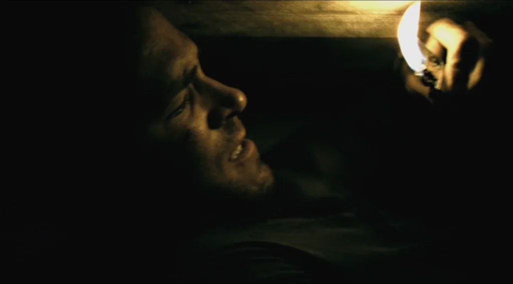 Buried (2010) movie still: closeup — A dark, cramped wooden space with extremely limited vertical clearance and no visible exits. A man lies on his…