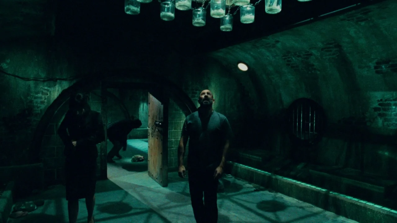 Saw V (2008) movie still: fullbody — A subterranean bunker with arched stone ceilings and a heavy iron door. A man stands in the foreground looking…