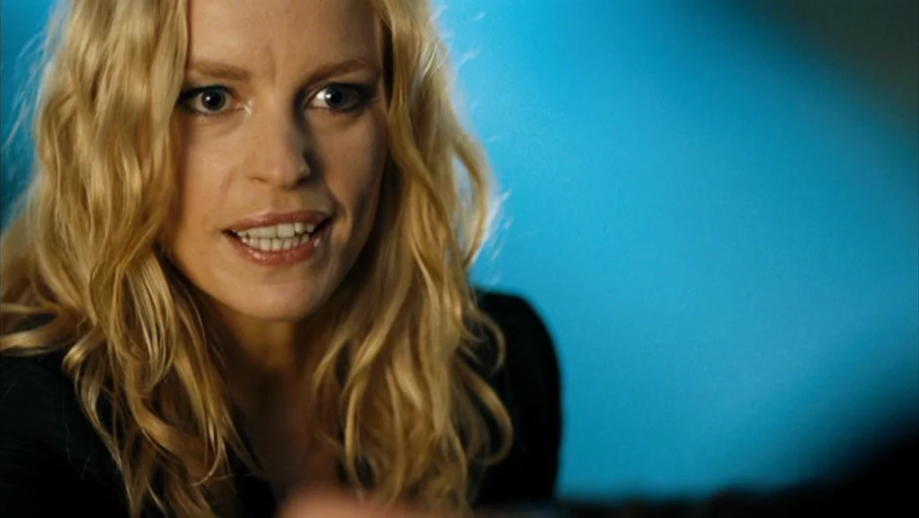 We Are The Night (2010) movie still: closeup — A close-up shot of a blonde woman standing in front of a blue wall. She speaks directly forward with…