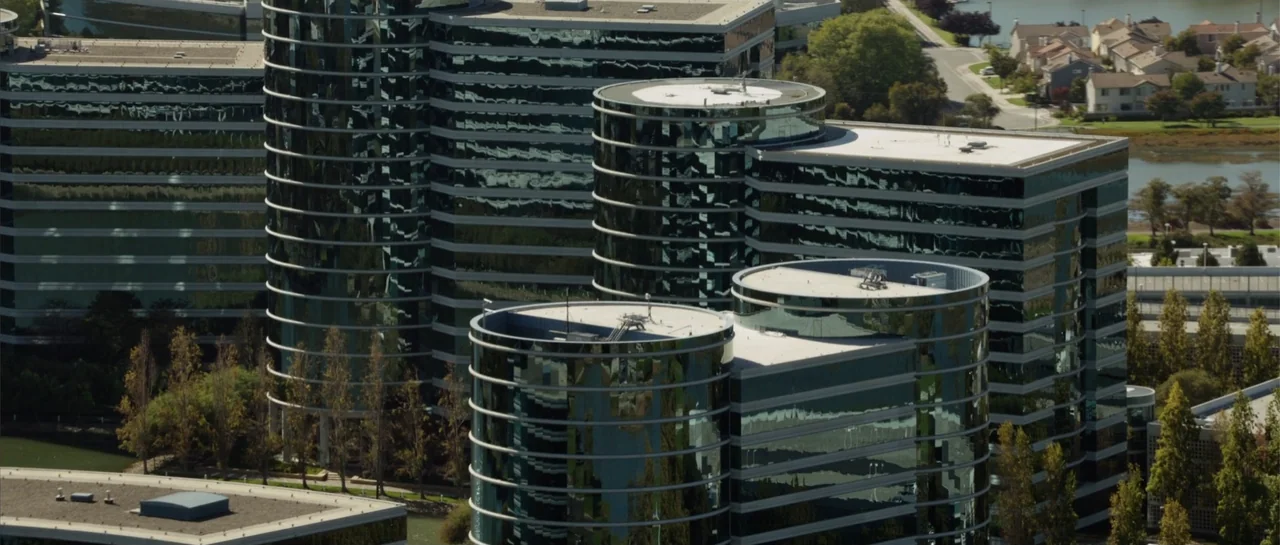 The Internship (2013) movie still: establishing — A cluster of modern glass office buildings with circular architecture stands in a park. Green…