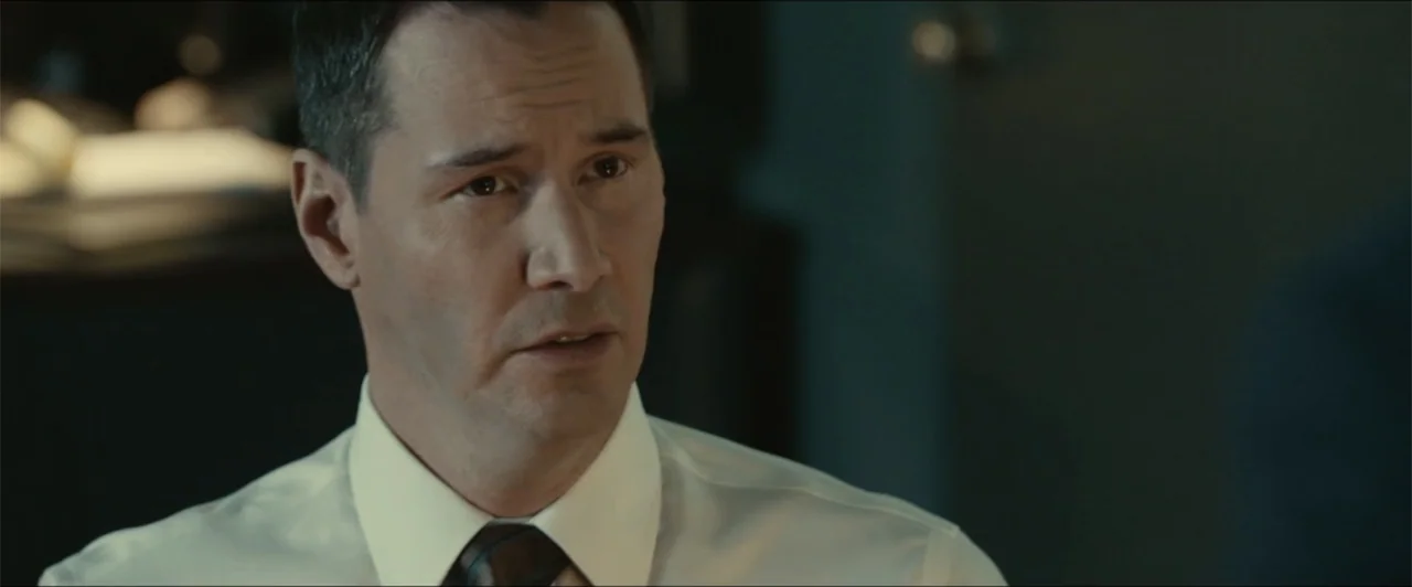 Exposed (2016) movie still: closeup — An interior office space with a blurred dark wall and ambient desk equipment. A middle-aged man in a dress…