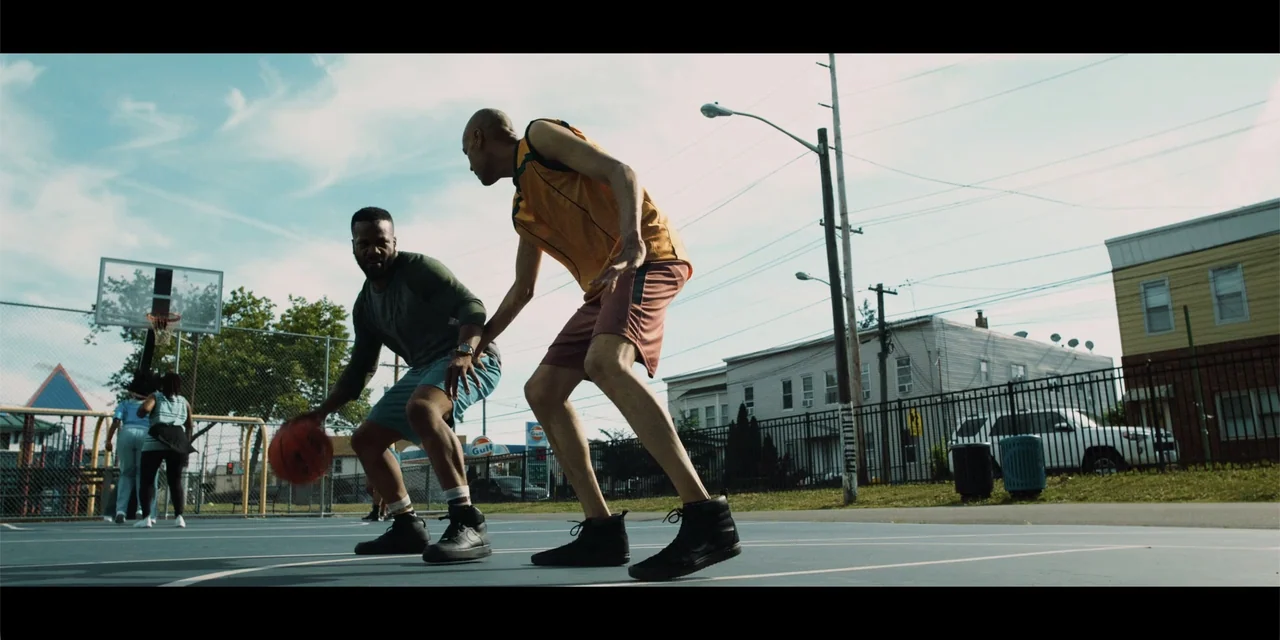 Música (2024) movie still: fullbody — An outdoor basketball court features a blue surface, chain-link fences, and surrounding residential buildings.…