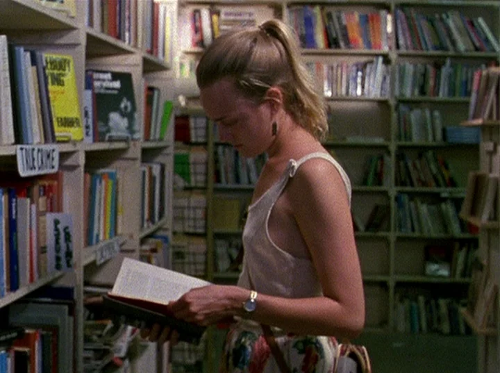 Slacker (1991) movie still: medium — Rows of white bookshelves filled with various books line a narrow aisle. A young woman stands in the aisle…