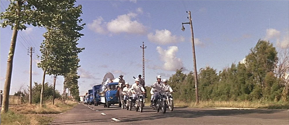 Les Demoiselles de Rochefort (1967) movie still: wide — A rural paved road lined with trees and utility poles under a clear sky. A group of men on…