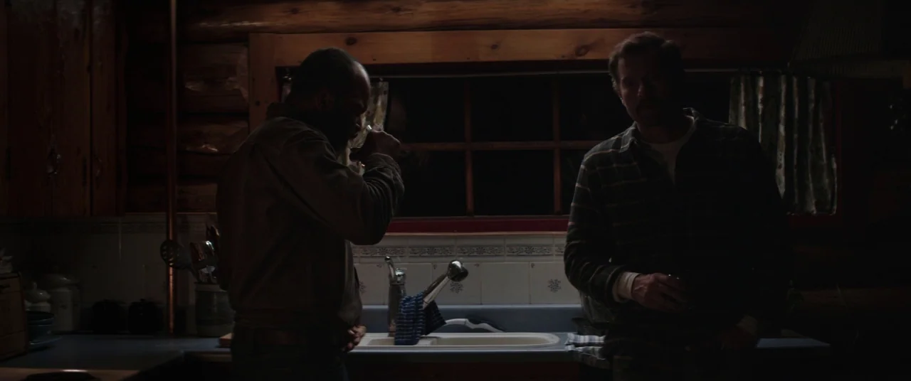 Hold The Dark (2018) movie still: medium — A dark, wood-paneled cabin interior features a sink and cabinets at night. Two men stand in the dim room…