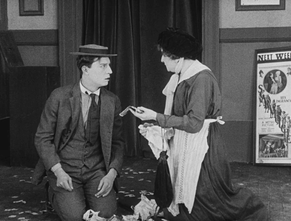 Sherlock Jr. (1924) movie still: medium — A theater lobby with a movie poster displayed on the wall near a doorway. A young man kneels on the floor…
