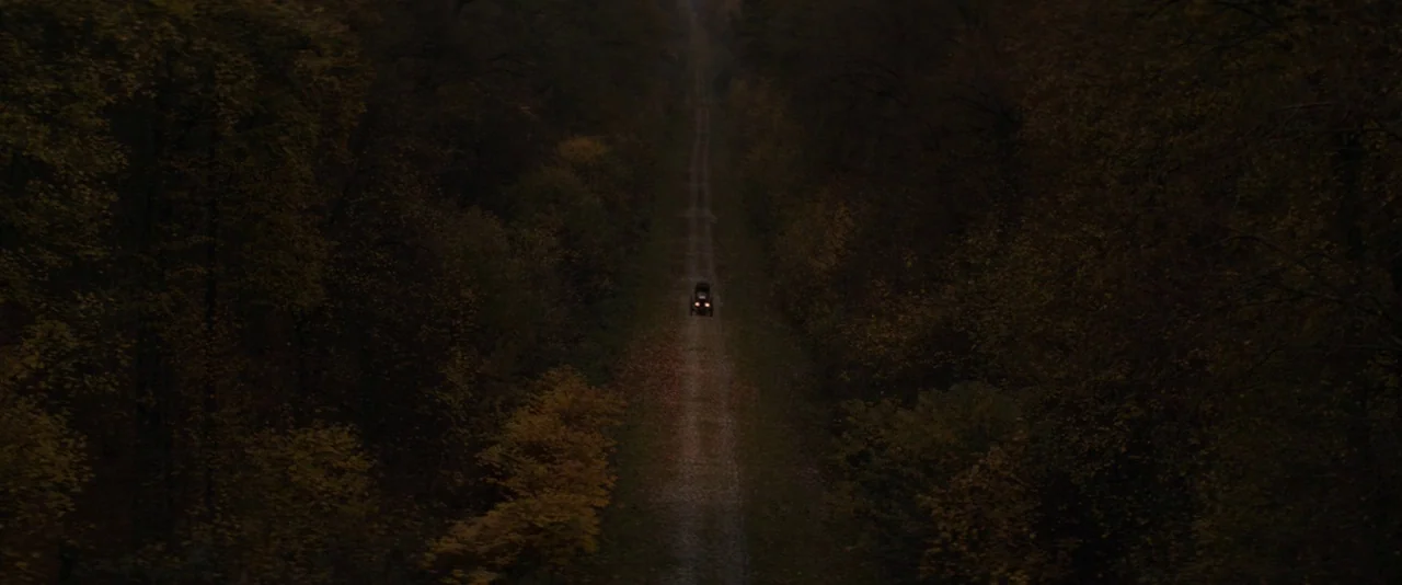 The Dancer movie still: establishing — A narrow dirt road stretches through a dense forest filled with dark autumn trees. A small vehicle with…