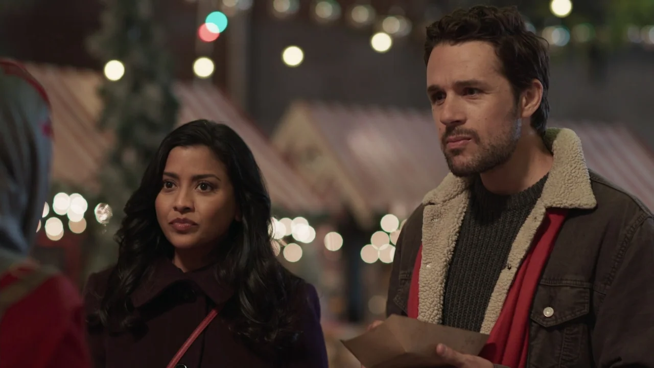 A Gingerbread Christmas (2022) movie still: medium — Two people stand before stalls decorated for the holiday season at night. The man holds a paper…