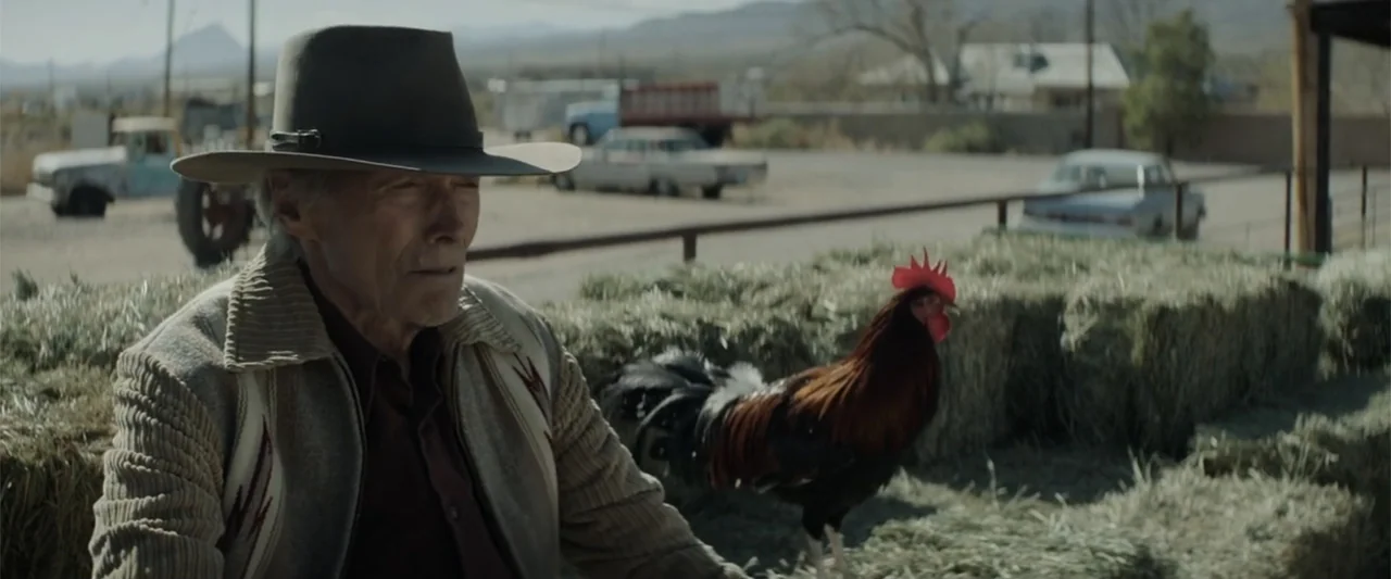 Cry Macho (2021) movie still: medium — A rural setting with bales of hay and old vehicles parked in the background. An elderly man in a hat sits…