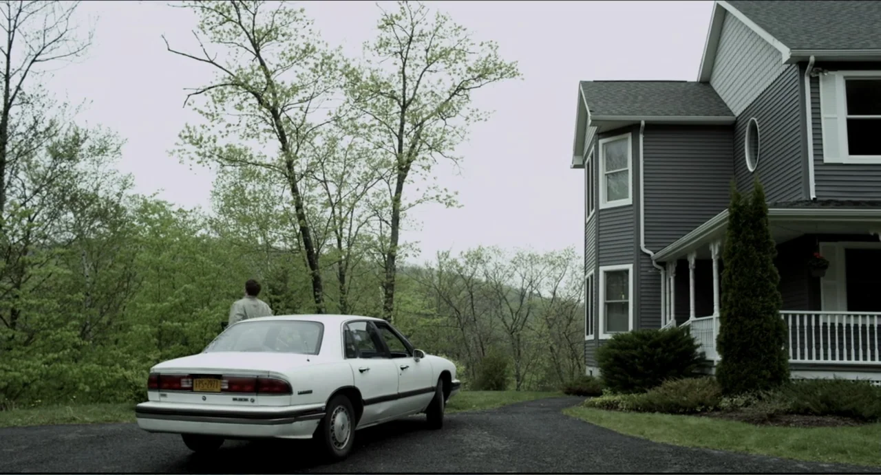 The Secret Village (2013) movie still: wide — A suburban house with grey siding sits next to a paved driveway and trees. A man in a tan jacket…