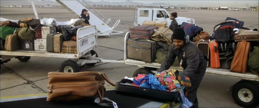 Three Kings (1999) movie still: medium — A flat asphalt airport tarmac features several metal carts stacked with various travel suitcases. An…