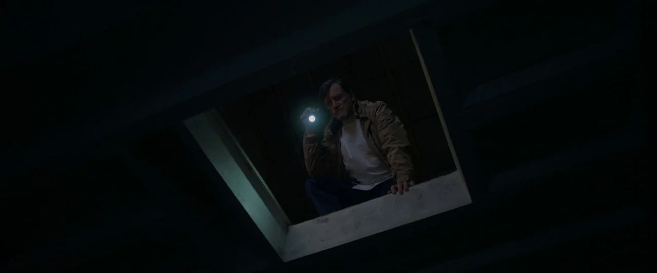 Oddity (2024) movie still: medium — A dark space with a rectangular opening looking down into a larger room. A middle-aged man peers through the…