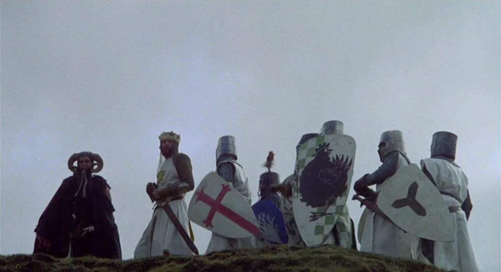 Monty Python and the Holy Grail (1975) movie still: fullbody — A group of medieval knights and a robed figure stand on a grassy knoll. An overcast…