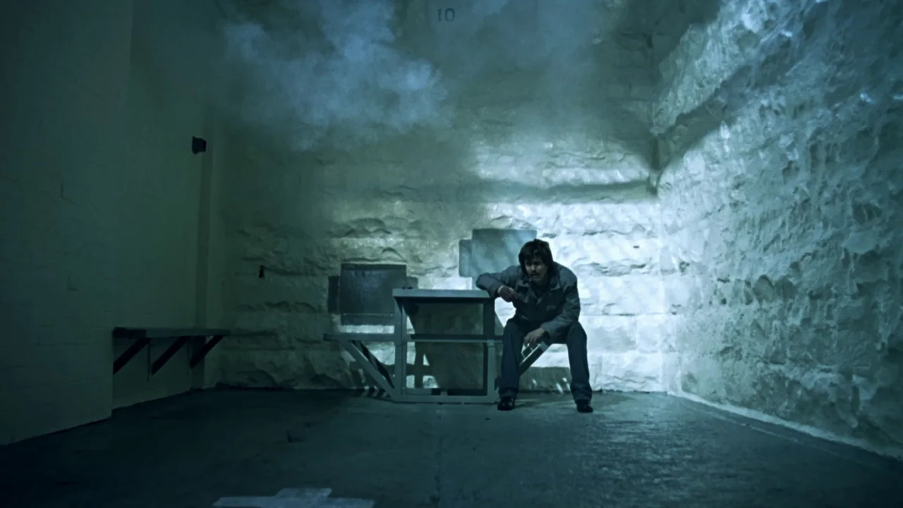 Chopper (2000) movie still: wide — A spartan prison cell features rough-hewn stone walls and a small metal table. A lone man sits on a bench while…
