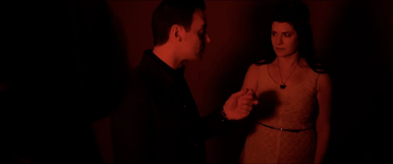 Amityville Cult (2021) movie still: medium — A dark indoor space is filled with intense red light. A man reaches his hand toward a woman in a…