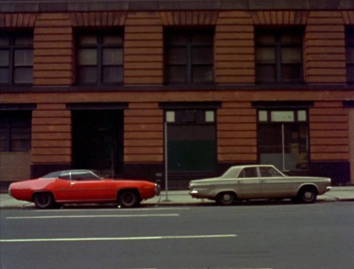 News From Home (1977) movie still: wide — A low-rise urban brick building features dark windows and recessed entrance alcoves. Two vintage…