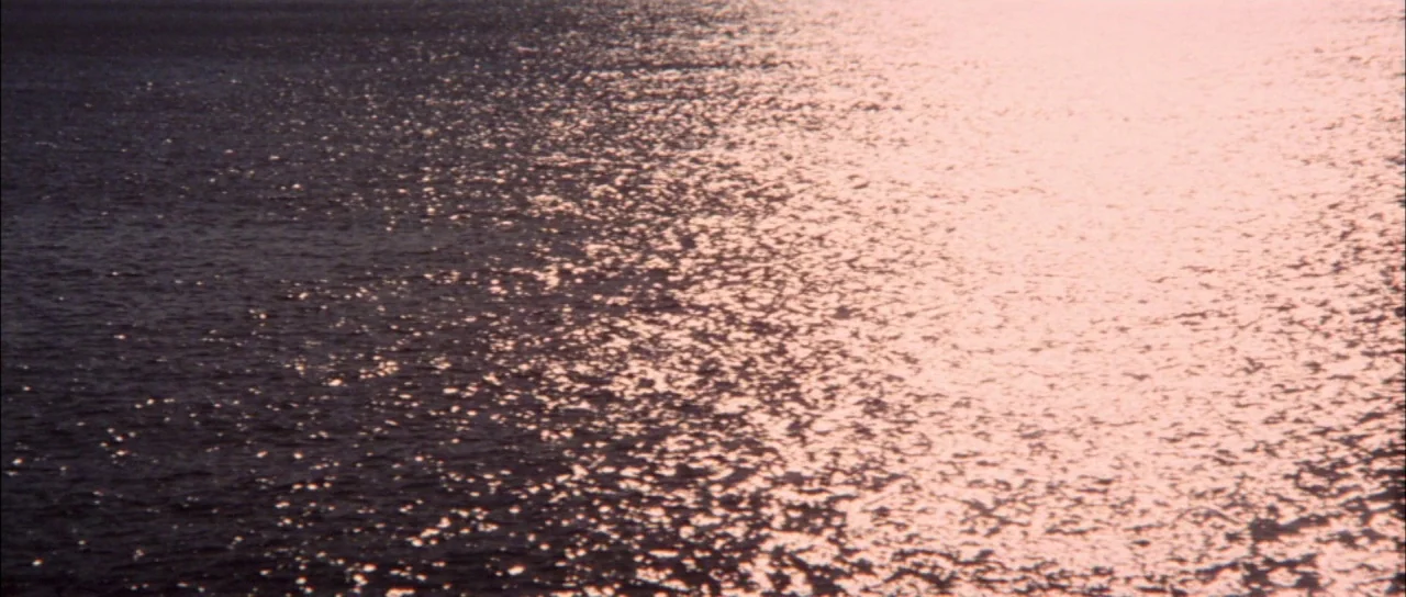 Terror of Mechagodzilla (1975) movie still: establishing — A vast expanse of water ripples under the golden light of the late afternoon. The surface…