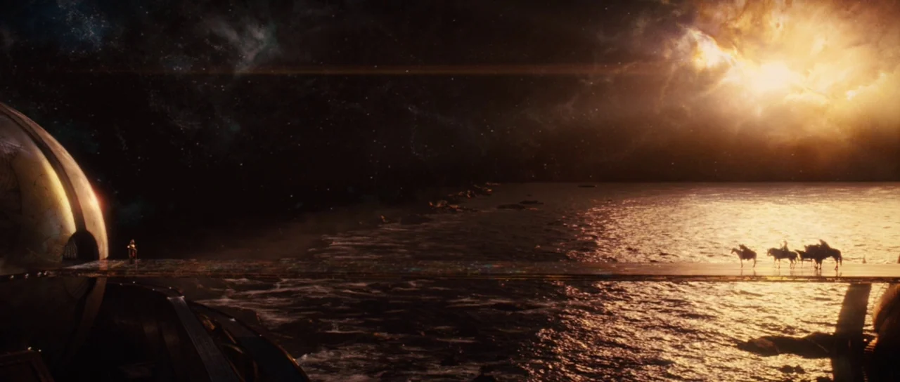Thor (2011) movie still: wide — A long, flat bridge extends over a dark body of water toward a massive structure. Riders on horseback travel across…