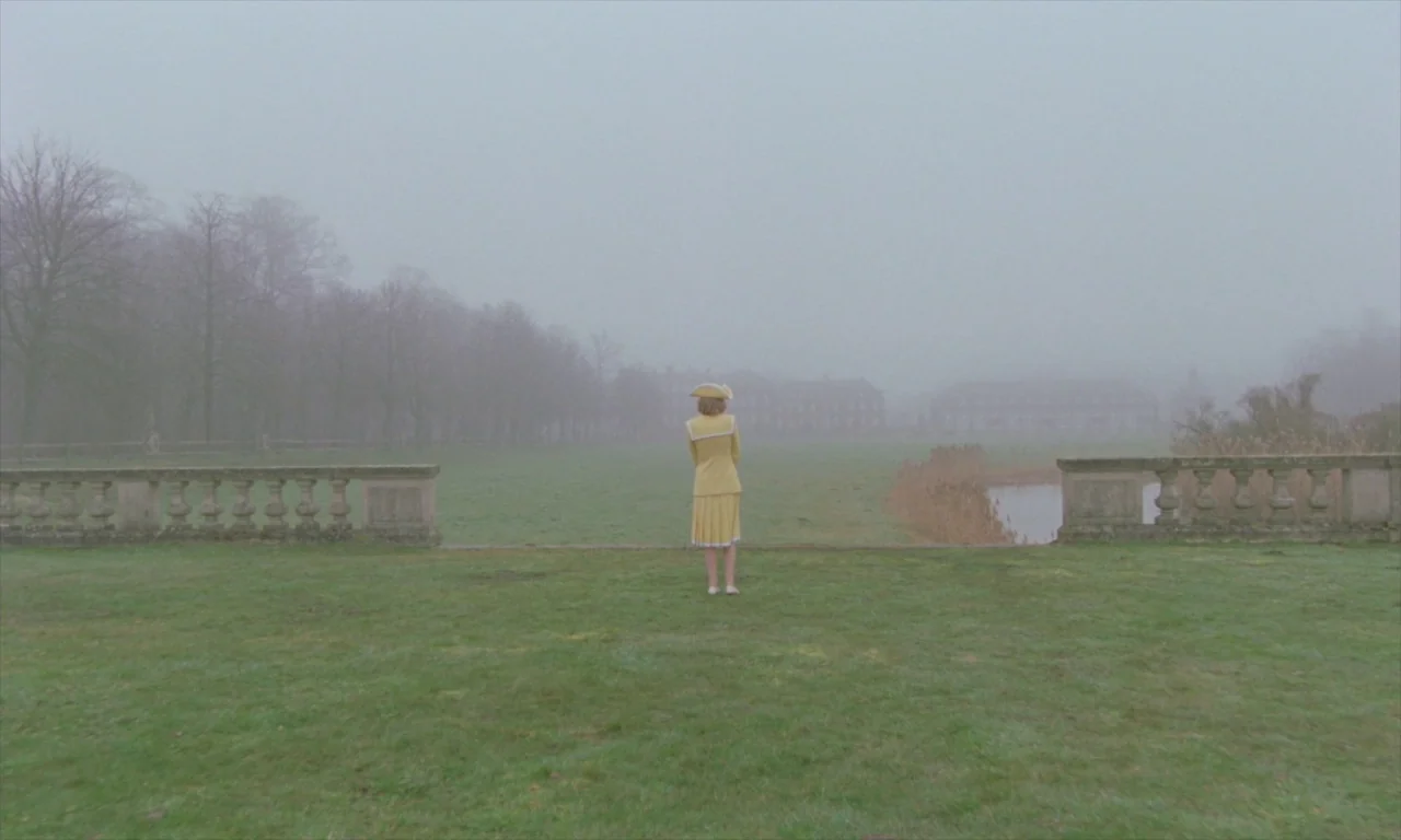 Spencer (2021) movie still: fullbody — A flat, grassy field stretches toward a distant building under a thick, grey fog. A woman in a bright yellow…