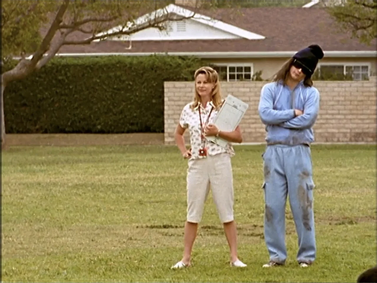 Just 4 Kicks (2003) movie still: fullbody — A green suburban lawn sits in front of a residential house with a brick wall. A woman holding a…