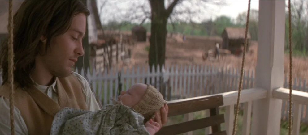 Ride With The Devil movie still: medium — A wooden porch with a white picket fence overlooks a rural farm setting. A man sits on a porch swing…