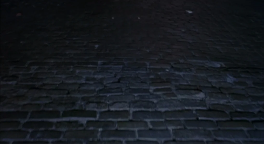 Light Sleeper (1992) movie still: establishing — A dark, worn cobblestone surface stretches across the frame in low-light conditions. No people or…
