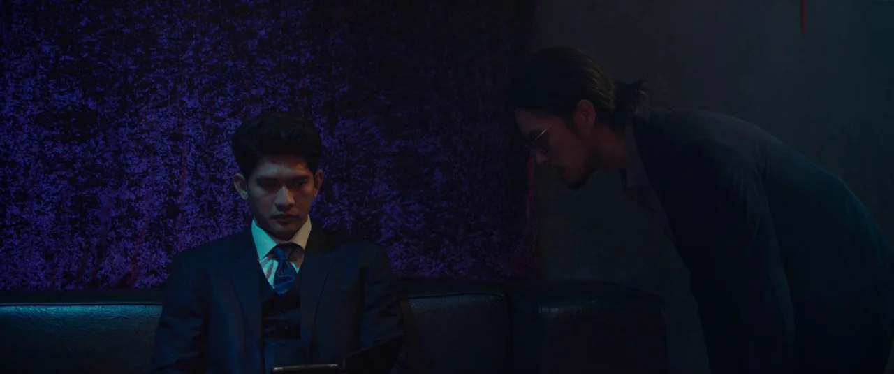 The Night Comes for Us (2018) movie still: medium — A dimly lit lounge with a dark textured purple wall as the background. Two men in formal…