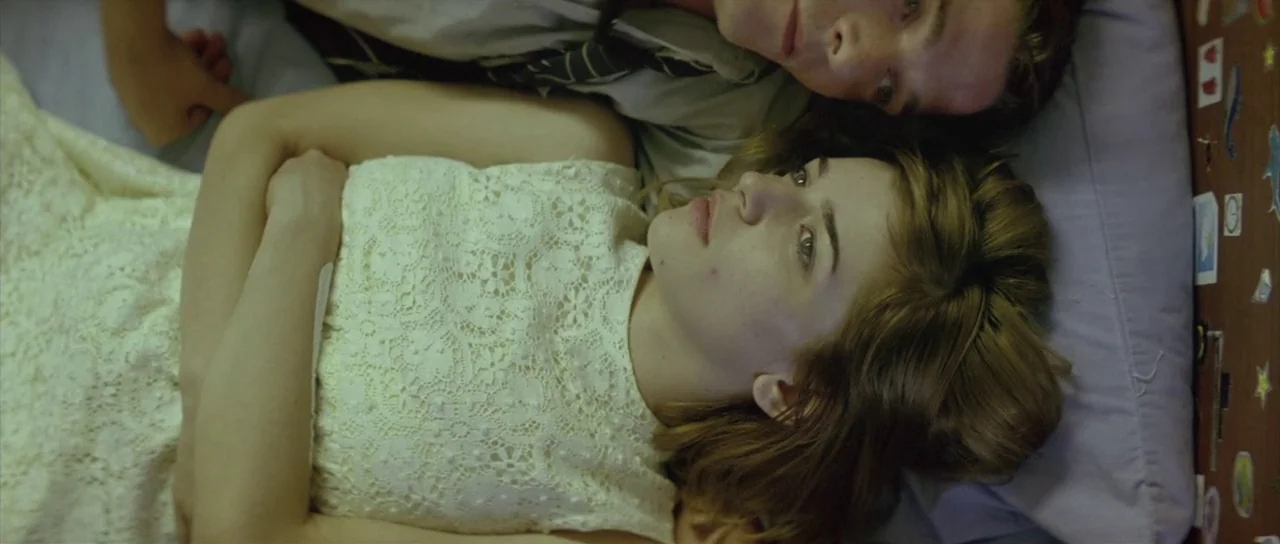 Dogtooth movie still: medium — A top-down view of two people lying on a mattress with a lavender pillow. The young woman in a lace top stares upward…