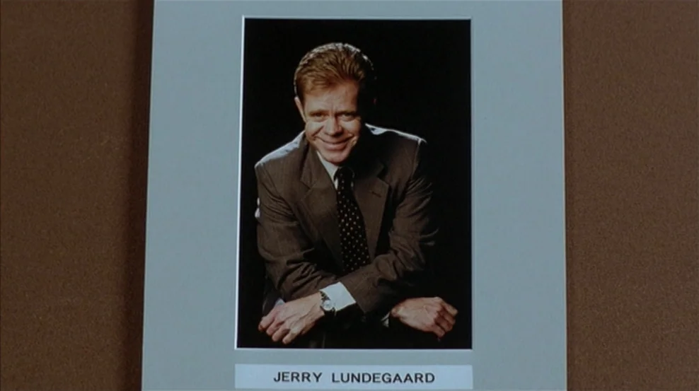 Fargo (1996) movie still: closeup — A framed professional portrait photograph of a man sits on a brown surface. The man in the photograph leans…