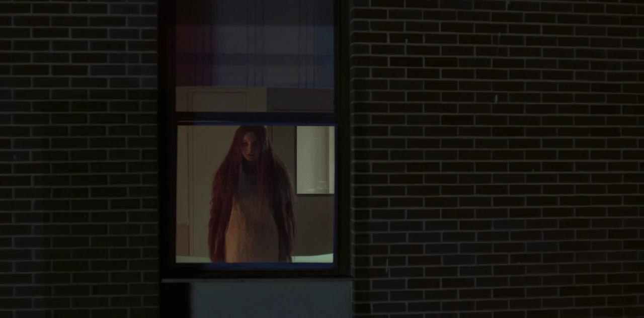 Darlin’ (2019) movie still: medium — A dark brick exterior wall surrounds a single illuminated window looking into a room. A woman stands in the…