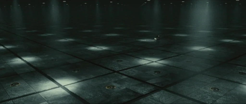 Sunshine movie still: establishing — A vast, dark industrial space features a floor composed of large, uniform metallic tiles. Rows of small,…
