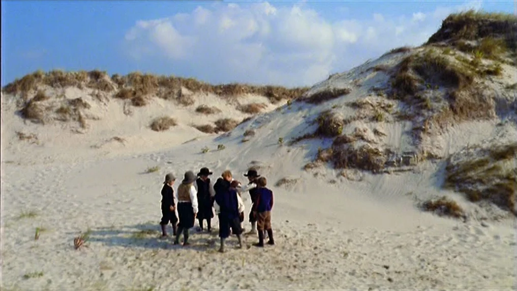 The Scarlet Letter (1973) movie still: wide — A vast, hilly landscape composed of light-colored sand dunes under a bright sky. A group of seven…