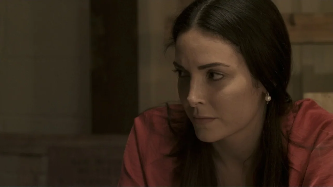 Locas y atrapadas (2014) movie still: closeup — A dimly lit interior space contains a person in front of a neutral backdrop. A woman with dark hair…