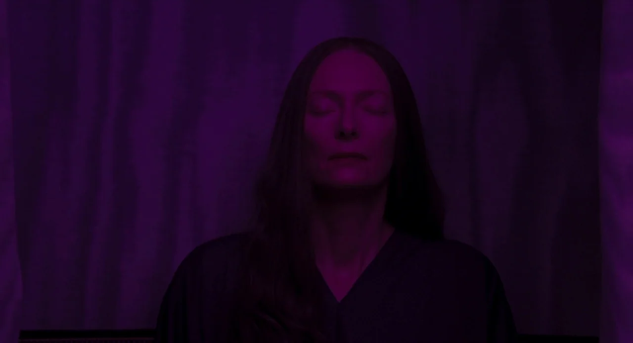 Suspiria (2018) movie still: closeup — A woman stands in a dimly lit room against a textured purple backdrop. She remains still with her eyes…