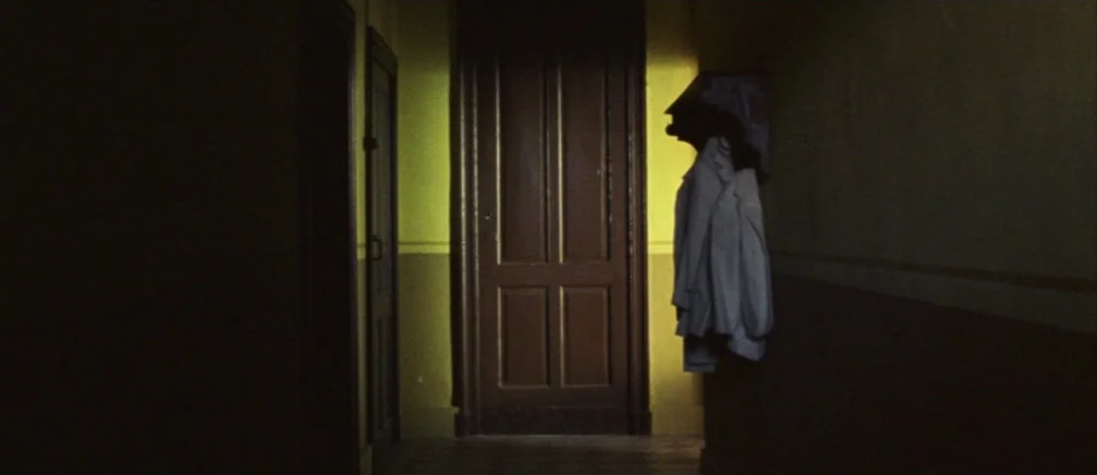 Innocence (2005) movie still: wide — An dimly lit interior hallway with yellow walls and dark wooden doors. A white coat hangs prominently on a hook…