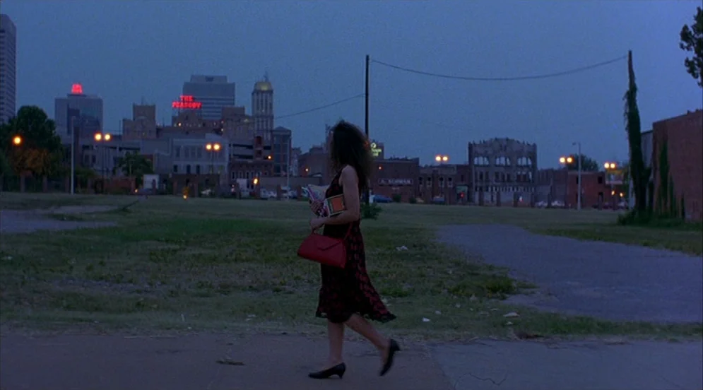 Mystery Train (1989) movie still: fullbody — A dark, grassy vacant lot stretches out before a distant, illuminated city skyline. A woman walks…