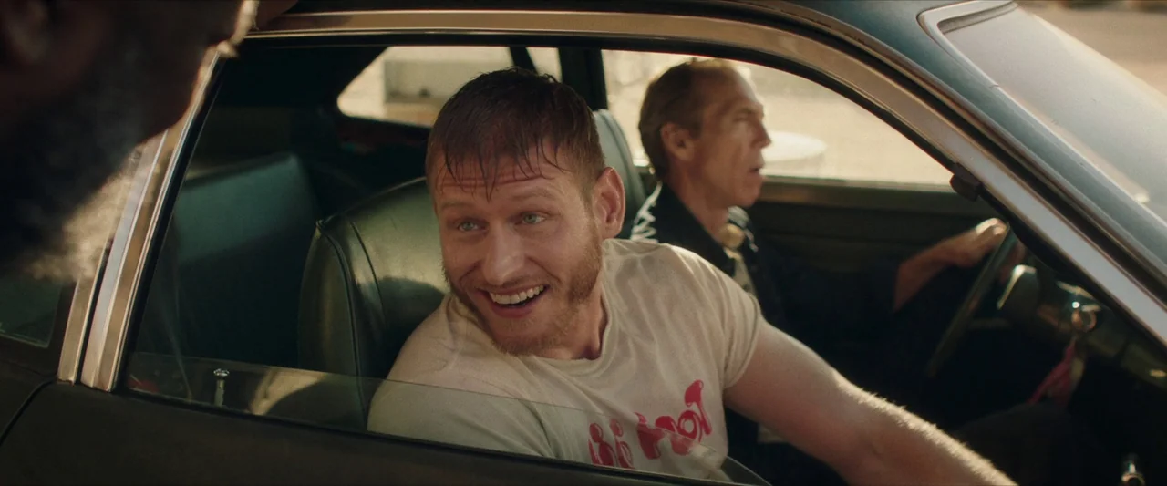 The Last Stop in Yuma County (2023) movie still: medium — A man in a graphic t-shirt sits in the passenger seat of a car. Beside him, an older man…