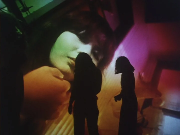 Butterfly (1974) movie still: medium — A dimly lit space features a large projection of a woman's face on a wall. Two silhouetted figures stand in…