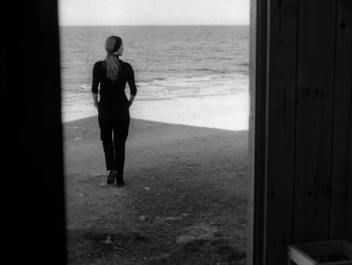 Persona (1966) movie still: fullbody — A woman walks on a pebbled beach toward the ocean seen from a doorway. She moves away from a wooden cabin…