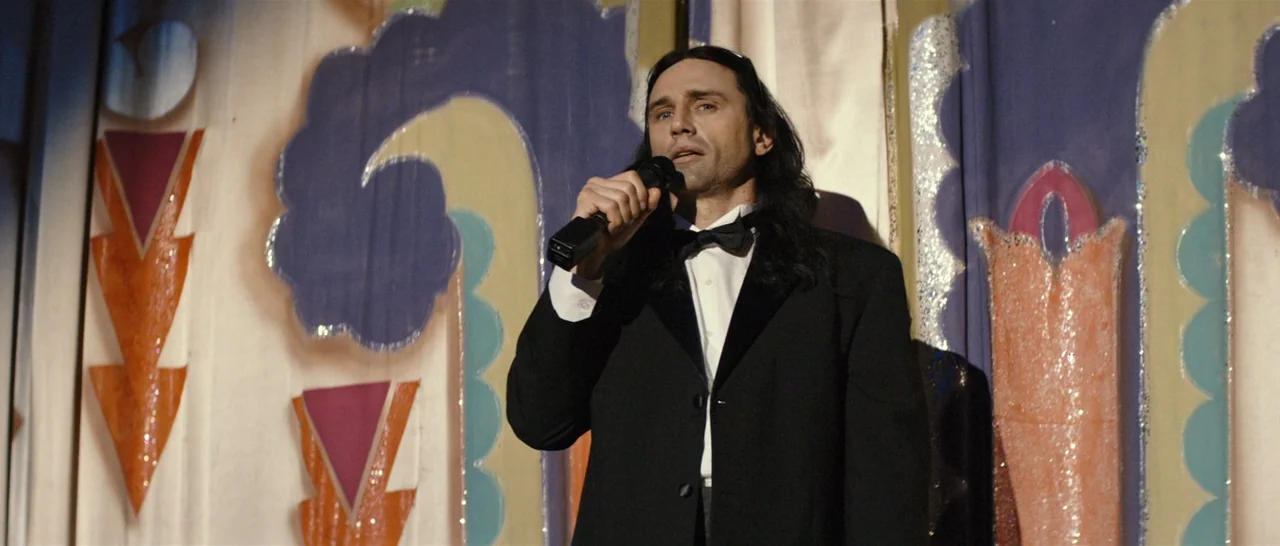 The Disaster Artist (2017) movie still: medium — A man in formal wear stands before a colorful, hand-painted curtain background. He holds a black…