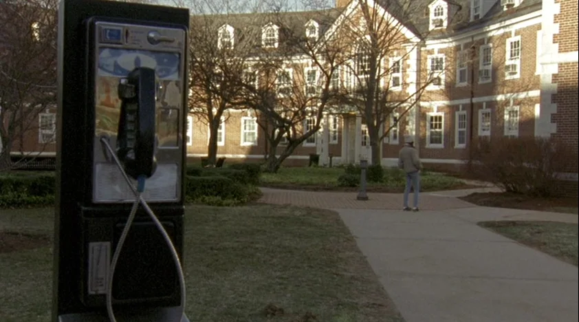 The House Of The Devil (2009) movie still: wide — A quiet campus courtyard features a red brick building with white window frames. A lone person…