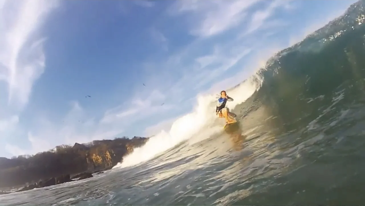 El sueño (2020) movie still: wide — A surfer rides a curling wave on a clear day with rocky cliffs nearby. The surfer balances on their board while…
