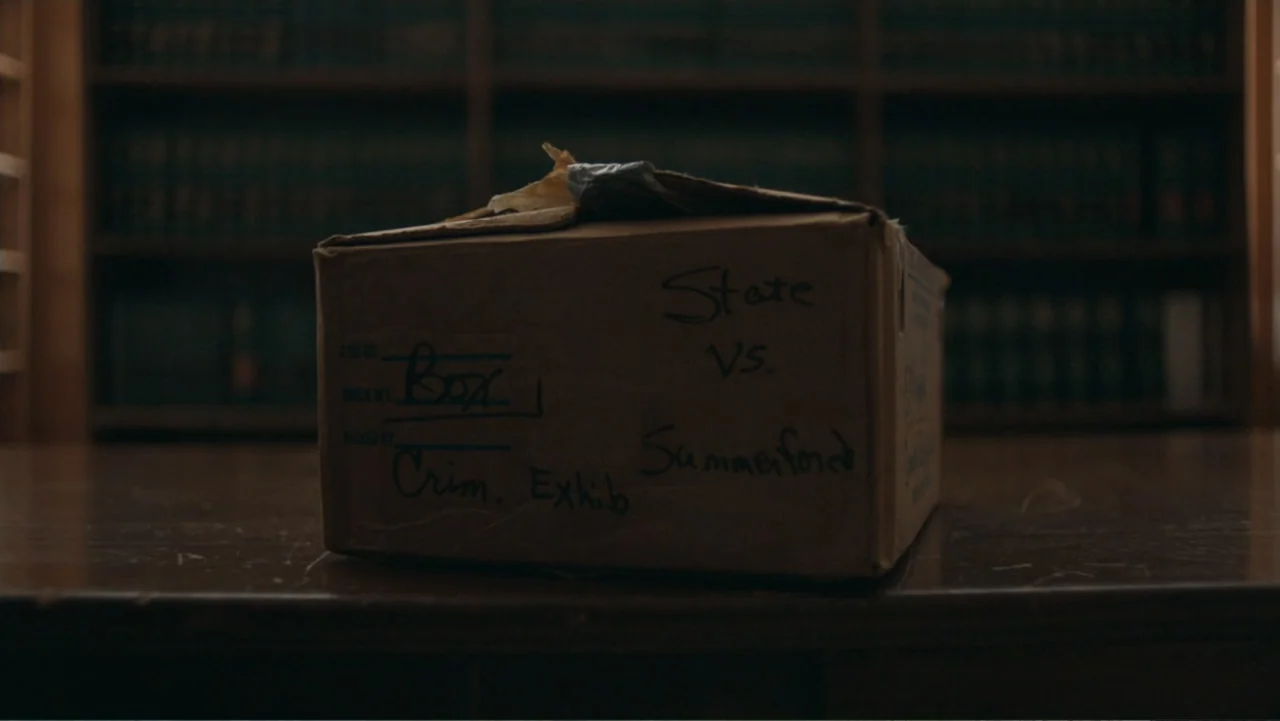 Alabama Snake (2020) movie still: closeup — A brown cardboard box labeled 'Crim. Exhib' sits on a dark wooden table. A blurry wall of bookshelves…
