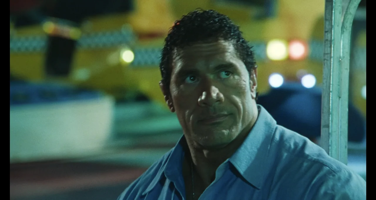 The Smashing Machine (2025) movie still: closeup — An exterior city street at night with a blurred yellow taxi in the background. A muscular man in…