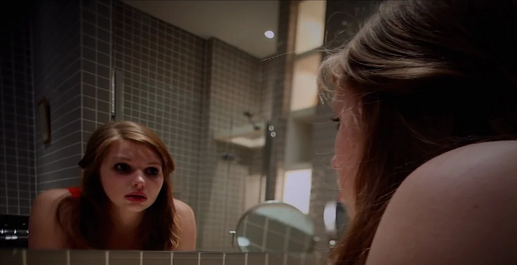 Dollhouse (2012) movie still: medium — A modern tiled bathroom features a large wall-mounted mirror above a counter surface. A young woman views her…