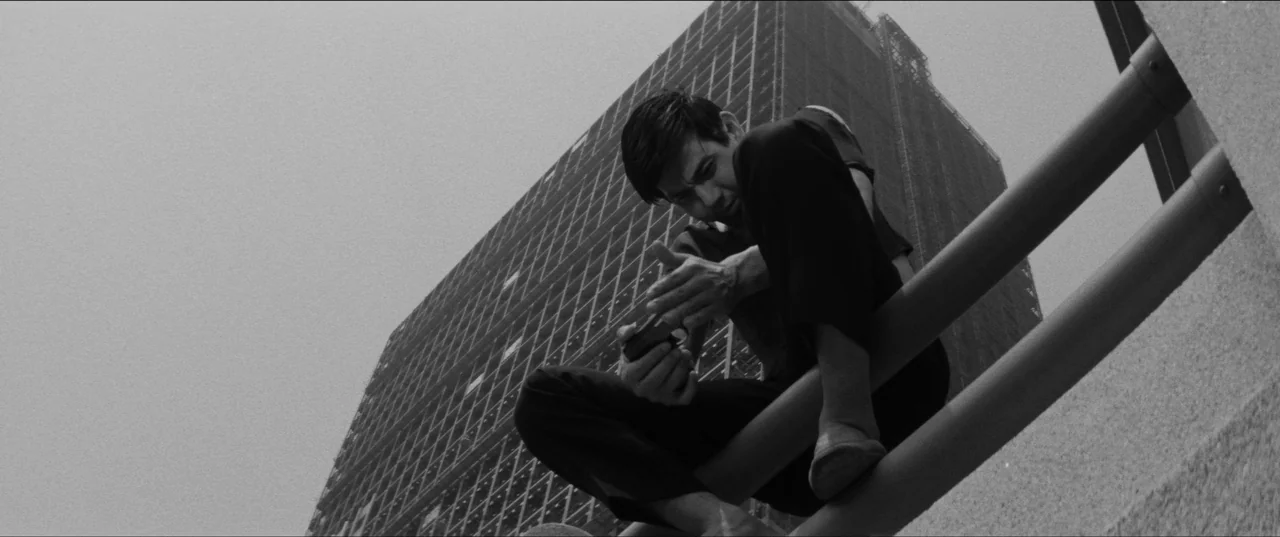 Branded To Kill (1967) movie still: medium — A man sits perched on a metal railing against a large, angular modern building. He looks down at a…