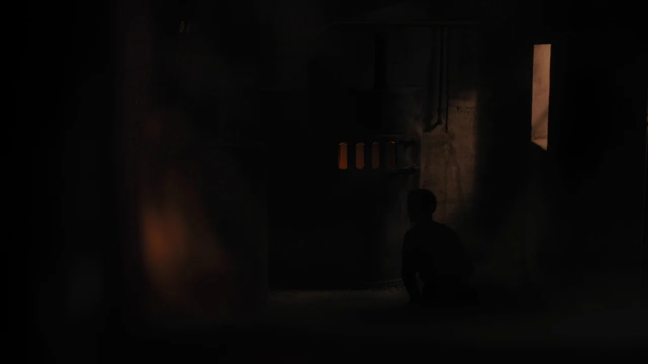 The Blackcoat’s Daughter (2015) movie still: wide — A dark, sparse basement features a large industrial metal furnace against a cinderblock wall. A…