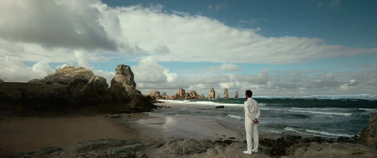 Oxygen (2021) movie still: wide — A rugged coastline features dramatic rock formations scattered along a sandy beach. A man wearing white stands on…